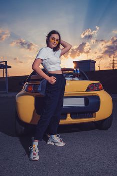 Attractive young girl in sunglasses, white top, black pants and sneakers is posing outdoors near yellow car cabriolet with two bottles of soda on its trunk. Summer sunset. Full length, copy space