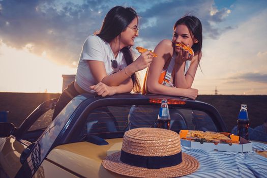 Two young girls in casual outfit eating pizza, laughing, posing in yellow car with french fries, hat and soda in glass bottles on trunk. Fast food. Summer sunset, cloudy sky. Close up, mock up