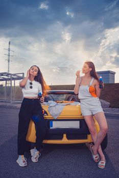 Gorgeous women in casual outfit are enjoying soda in glass bottles and eating french fries, posing leaning on trunk of yellow car with pizza on it. Fast food. Parking lot. Full length, mock up