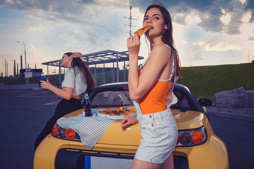 Attractive women in casual clothes are eating pizza, enjoying carbonated drink in glass bottles while posing near yellow car on parking lot. Fast food. Summer evening. Close up, copy space, mock up
