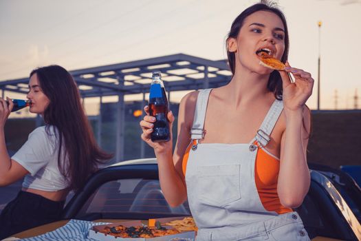 Attractive girls in casual outfit are eating pizza, enjoying carbonated drink in glass bottles while posing near yellow car on parking lot. Fast food. Summer evening. Close up, copy space, mock up