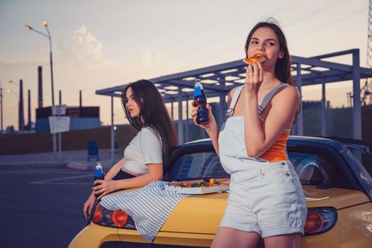 Attractive ladies in casual clothes are eating pizza, enjoying soda water in glass bottles while posing near yellow car on parking lot. Fast food. Summer evening. Close up, copy space, mock up