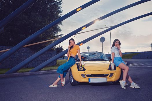 Gorgeous girls in casual outfit are eating hamburgers and holding beverages in paper cups while leaning on the hood of yellow car roadster. Fast food. Sunny day, iron pipes arch. Copy space, mock up