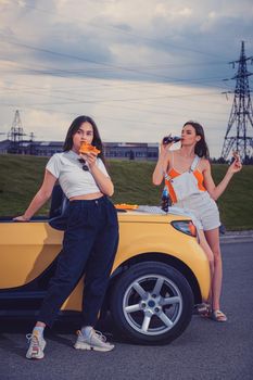 Attractive models in casual outfit are eating pizza, enjoying soda water in glass bottles while posing near yellow car on parking lot. Fast food. Summer evening. Full length, copy space, mock up