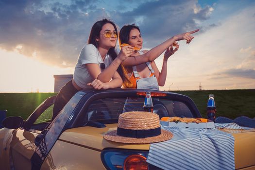 Charming models in casual clothes eating pizza, pointing at something, posing in yellow car with french fries and soda in glass bottles on trunk. Fast food. Summer sunrise, cloudy sky. Copy space