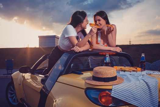 Two young females in casual clothes eating pizza, smiling, posing in yellow car with french fries, hat and soda in glass bottle on its trunk. Fast food. Summer sunrise, cloudy sky. Mock up, copy space