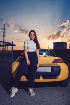 Beautiful young female in sunglasses, white top, blue jeans and sneakers is posing outdoors near yellow car roadster with two glass bottles of soda on its trunk. Summer sunset. Full length, mock up