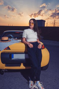 Attractive young lady in sunglasses, white top, blue jeans and sneakers is posing outdoors near yellow car roadster with pizza on its trunk. Summer sunset. Fast food. Full length