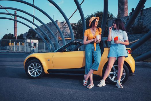 Young ladies in casual outfit are smiling, enjoying french fries from paper package and beverages in paper cups, posing near yellow car cabriolet. Fast food. Iron pipes arch. Copy space, mock up