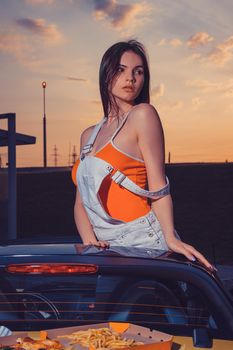 Sexy young woman in white overall and orange t-shirt is posing outdoors standing in yellow car cabrio with french fries and pizza on its trunk. Fast food, unhealthy nutrition. Summer sunset. Close up