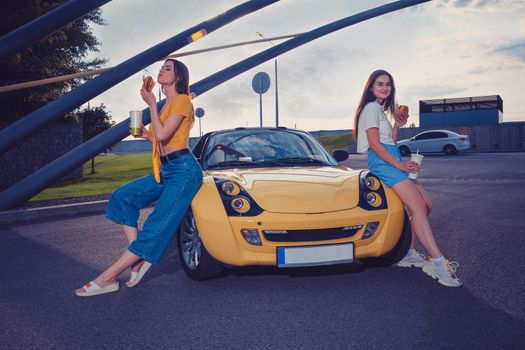 Gorgeous women in casual outfit are enjoying hamburgers, holding beverages in paper cups while sitting on the hood of yellow car. Fast food. Sunny day, industrial zone. Copy space, mock up