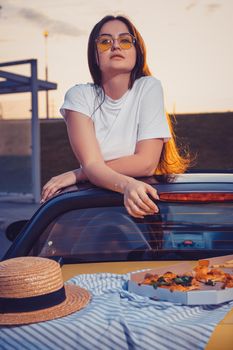 Young brunette lady in sunglasses, white t-shirt is posing in yellow car cabriolet with pizza, striped cloth and hat on its trunk. Fast food, unhealthy nutrition. Summer evening. Close up