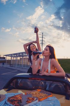 Happy young girls in casual outfit having fun and drinking soda in glass bottles, posing in yellow car cabrio with french fries and pizza on its trunk. Fast food. Summer evening. Copy space, mock up