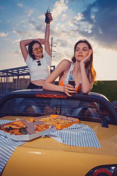 Happy ladies in casual clothes having fun, drinking soda water in glass bottles, posing in yellow car cabriolet with french fries and pizza on trunk. Fast food. Summer evening. Close up, copy space