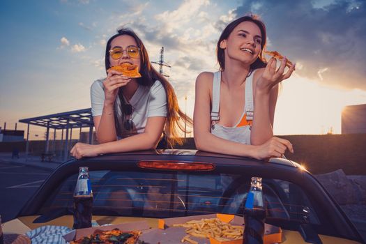 Charming women in casual clothes are eating pizza and smiling while posing in yellow car cabriolet with french fries and soda in glass bottles on trunk. Fast food. Summer evening. Close up, copy space