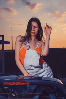 Sexy young female in white overall and orange t-shirt is posing outdoors standing in car cabriolet. Summer evening, sunset. Beauty, fashion, transportation. Close up