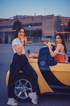 Pretty females in casual clothes are eating pizza, posing in yellow car with french fries and soda in glass bottle on its trunk. Fast food. Industrial zone. Summer evening. Full length, copy space
