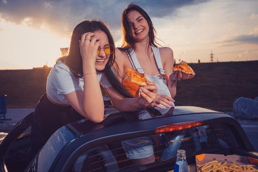 Two young ladies in casual clothes eating pizza, laughing, posing in yellow car with french fries and soda in glass bottle on trunk. Fast food. Summer sunset, cloudy sky. Close up, mock up