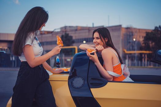 Pretty young girls in casual outfit are smiling, eating pizza, posing in yellow car cabrio with french fries and soda in glass bottle on its trunk. Fast food. Industrial zone. Close up, copy space