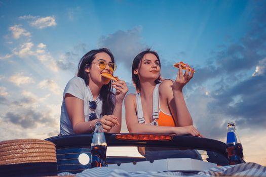 Two lovely women in casual clothes are eating pizza while posing in car cabrio with hat and soda in glass bottles on its trunk. Fast food. Summer sunny day, blue sky. Close up, copy space, mock up