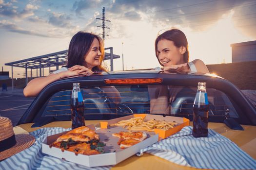 Young females in casual outfit are having fun, posing in yellow car roadster with french fries, pizza and soda in glass bottles on its trunk. Fast food. Summer evening. Close up, copy space, mock up