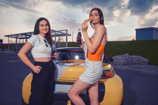Gorgeous friends females in casual outfit are smiling, eating pizza, enjoying soda in glass bottles while posing near yellow car on parking. Fast food. Summer evening. Close up, copy space, mock up