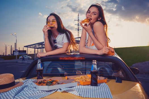 Charming females in casual outfit are eating pizza while posing in yellow car cabriolet with french fries and soda in glass bottles on trunk. Fast food. Summer evening, bus stop. Close up, copy space