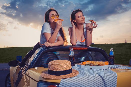 Charming young ladies in casual outfit are eating pizza, posing in yellow car cabrio with french fries, hat and soda in glass bottles on trunk. Fast food. Summer sunset, cloudy sky. Close up, mock up