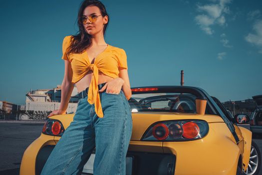 Young brunette girl in blue jeans, orange top and sunglasses is posing leaning on yellow car cabriolet with paper cup of beverage on trunk. Sunny day at parking lot. Close up, copy space, mock up