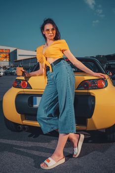 Young brunette model in blue jeans, orange top and sunglasses. She is holding paper cup of coffee, posing leaning on yellow car cabriolet at parking lot. Sunny day. Full length, copy space, mock up