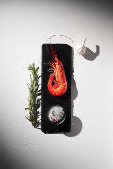 One cooked royal red shrimp on black slate board with coarse sea salt and peppercorns in saltcellar garnished with garlic bulb and fresh rosemary.Top view on grey background with shadow and copyspace