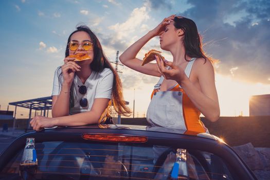 Happy young models in casual outfit are eating pizza while posing in yellow car roadster with soda water in glass bottles on its trunk. Fast food. Summer evening. Close up, copy space, mock up