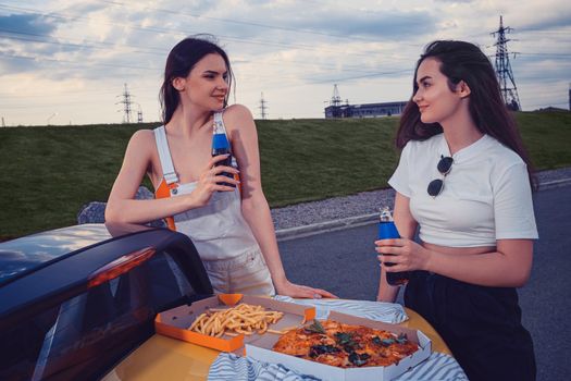 Young models in casual clothes are smiling, enjoying soda in glass bottles, posing leaning on trunk of yellow car with pizza and french fries on it. Fast food. Car parking. Close up, copy space