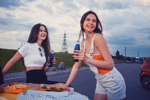 Young ladies in casual outfit are smiling, enjoying soda in glass bottles, posing leaning on trunk of yellow car with pizza and french fries on it. Fast food. Parking lot. Close up, mock up