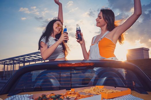 Two lovely young girls in casual clothes smiling, cheering with soda in glass bottles, posing in yellow car cabrio with french fries and pizza on trunk. Fast food. Summer sunset. Close up, copy space