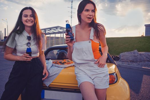 Gorgeous girls in casual outfit are laughing and enjoying soda in glass bottles while leaning on trunk of yellow car with pizza on it. Fast food. Parking lot, summer day. Close up, copy space, mock up