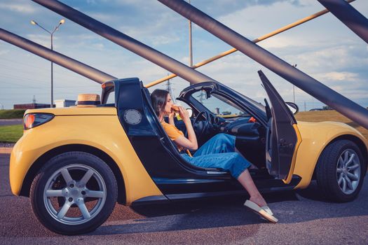 Attractive young girl in blue jeans, orange top and flip flops is eating hamburger while sitting in yellow car cabrio. Fast food, unhealthy nutrition. Sunny day, iron pipes arch. Side view