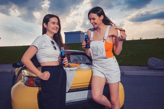 Gorgeous ladies in casual clothes are laughing, enjoying soda in glass bottles and pizza while leaning on trunk of yellow car. Fast food. Parking lot, summer day. Close up, copy space, mock up