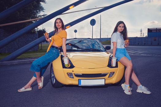 Gorgeous females in casual clothes are holding hamburgers and beverages in paper cups while sitting on the hood of yellow car cabriolet. Fast food. Industrial zone. Copy space, mock up