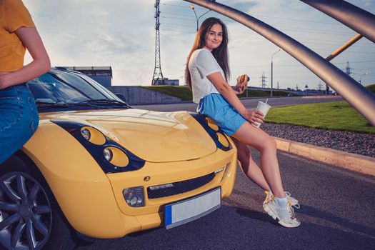 Attractive female in casual outfit is holding hamburger and drink in paper cup, sitting on the hood of yellow car cabrio with her friend. Fast food. Sunny day, iron pipes arch. Close up, copy space