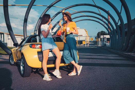 Young stylish women in casual clothes are smiling, enjoying french fries and beverages in paper cups while posing near yellow car cabriolet. Fast food. Sunny day, industrial zone. Copy space, mock up