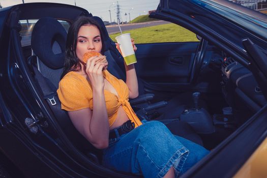 Attractive model in blue jeans and orange top is eating hamburger and holding beverage in paper cup while sitting in yellow car cabriolet. Fast food. Side view. Close up, copy space, mock up