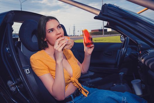 Beautiful female in blue jeans and orange top is eating hamburger and holding paper package with french fries while sitting in yellow car. Fast food. Side view. Close up, copy space, mock up