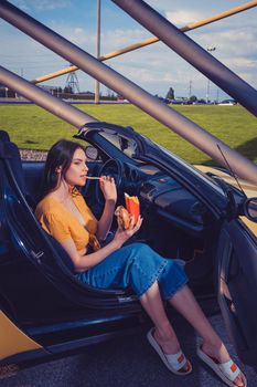 Beautiful woman in blue jeans and orange top is eating french fries from paper package and holding hamburger while sitting in yellow car cabrio. Fast food. Side view. Copy space, mock up