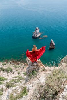 A girl with loose hair in a long red dress descends the stairs between the yellow rocks overlooking the sea. A rock can be seen in the sea. Sunny path on the sea from the rising sun.