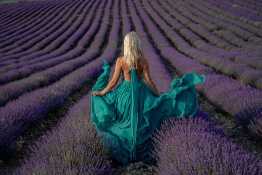 A woman with long blonde hair in a light green dress is walking through a lavender field alone waving a lavender dress