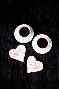 on a black background are two cups of coffee and biscuits in the form of heart.
