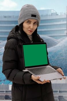 a brunette girl in a down jacket with a laptop in a gray hat stands against the background of the building