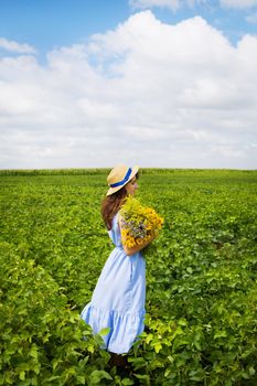 beautiful girl stands in the field with a bouquet of yellow flowers and a straw hat.