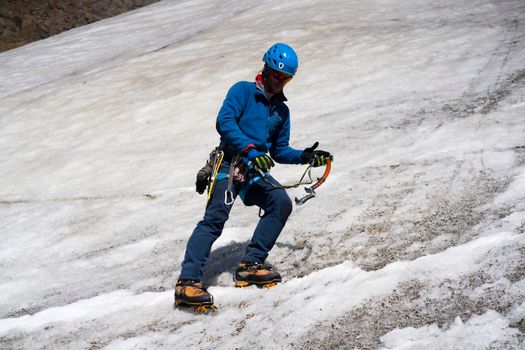Young stylish man in sunglasses, a traveler with ice tools while hiking in the mountains, a tourist with climbing equipment, in a helmet, crampons walks on a glacier, a snow-covered mountain.